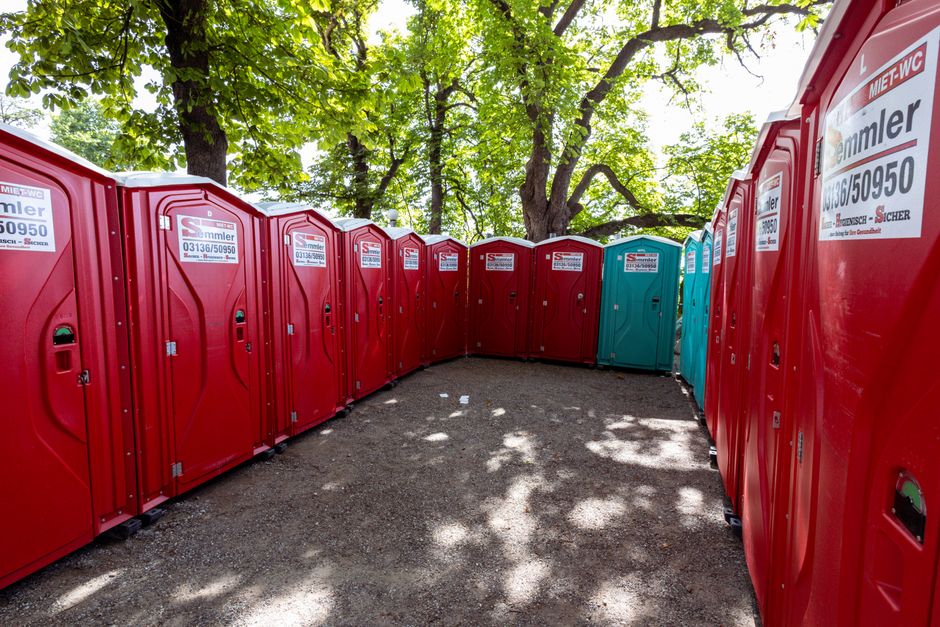 A row of red portable toilets lined up outdoors under green trees on a sunny day, ideal for outdoor event rental.